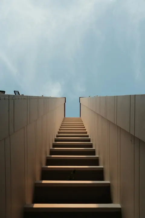 A minimalist staircase ascending between concrete walls toward an opening of blue sky.