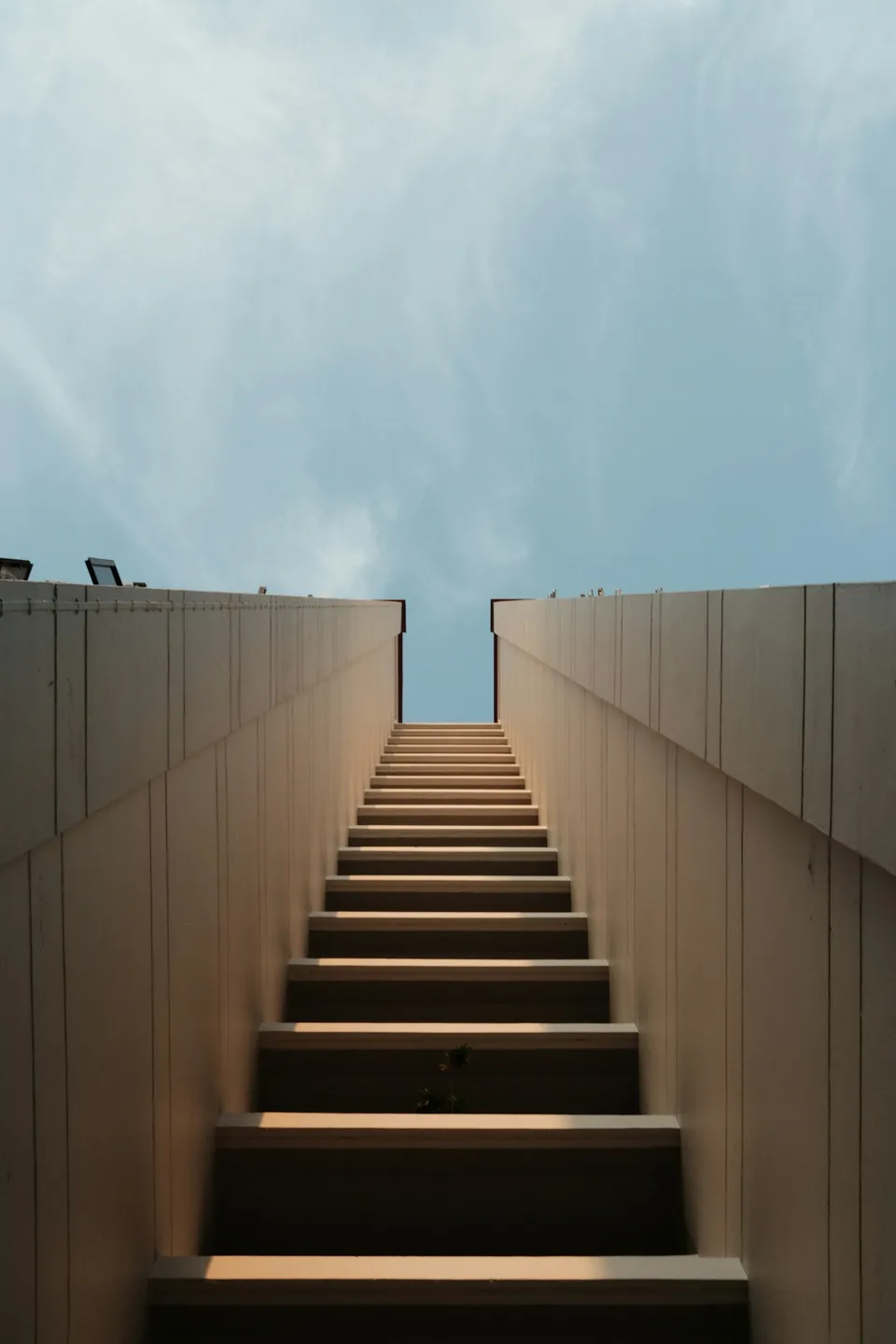 A minimalist staircase ascending between concrete walls toward an opening of blue sky.