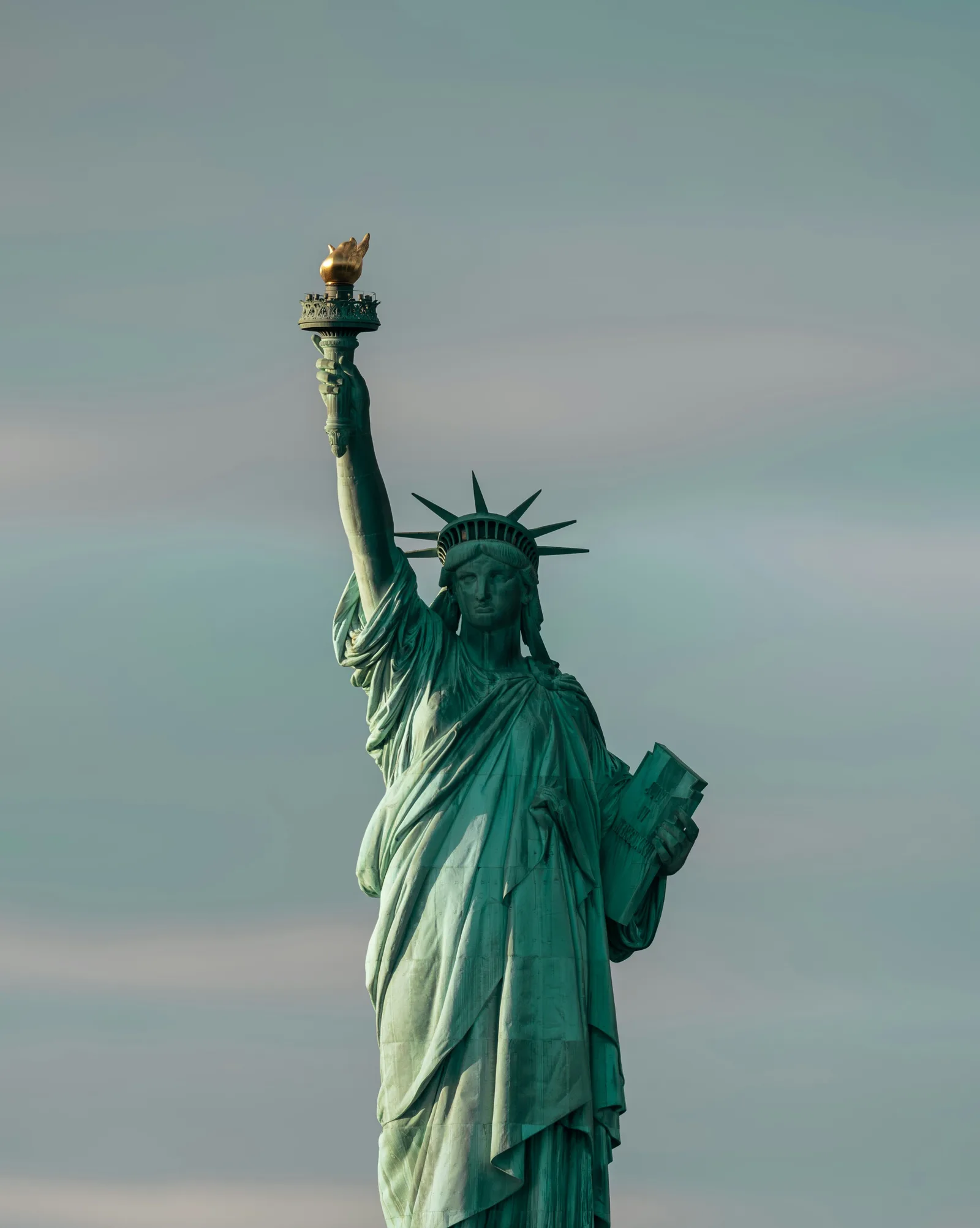 The Statue of Liberty photographed against a pale blue and gray sky, with its torch raised above New York Harbor.