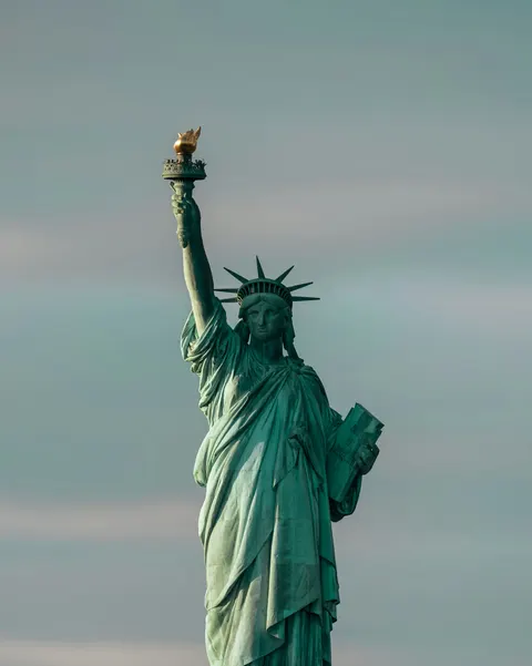 The Statue of Liberty photographed against a pale blue and gray sky, with its torch raised above New York Harbor.
